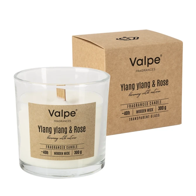 Świeca zapachowa z drewnianym knotem YLANG YLANG & RÓŻA sn100-000-384 Valpe ustawiona w eleganckiej aranżacji wnętrza z dekoracyjnymi dodatkami. Stylowa świeca w szkle podkreśla atmosferę relaksu i domowego komfortu.
