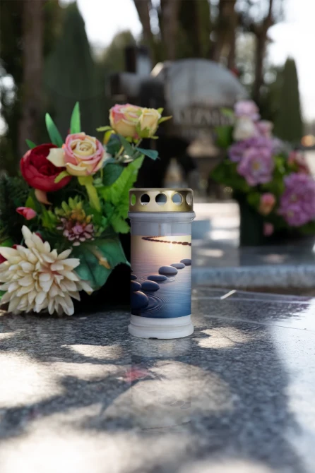 Znicz z motywem kamieni na wodzie ustawiony na grobie w otoczeniu kwiatów. Znicz zalewany o czasie palenia do 72 godzin, zapewniający stabilny płomień i estetyczny wygląd. Dekoracja nagrobna odpowiednia na cmentarz, święta i codzienne odwiedziny.