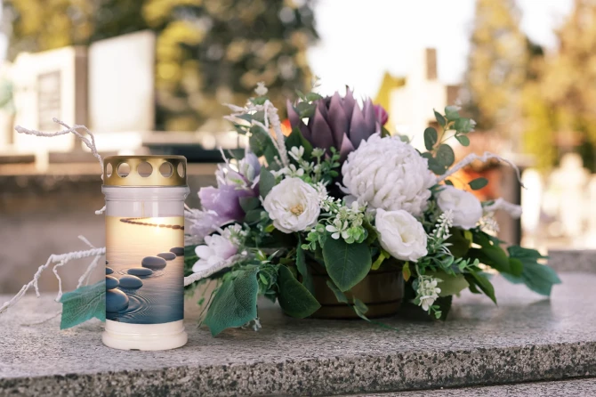 Znicz z motywem kamieni na wodzie w aranżacji nagrobnej z kwiatami. Znicz na grób o czasie palenia do 72 godzin, z metalowym wieczkiem chroniącym płomień przed wiatrem. Elegancki znicz nagrobny na Wszystkich Świętych, rocznice i inne dni pamięci.