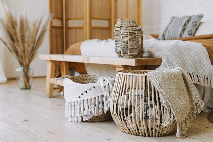 Linia zapachowa eco-design. Na zdjęciu widać przytulne pomieszczenie w stylu eco design. Dominuje drewno – drewniana podłoga i łóżko z naturalnego materiału. Na łóżku leży miękki koc w jasnym kolorze, a obok stoją plecione kosze do przechowywania. Całość jest jasna, naturalna i bardzo harmonijna.